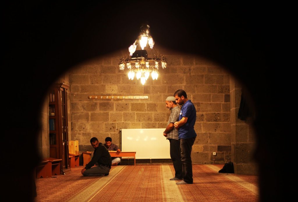 A serene scene of men praying inside a mosque in Erzurum, Türkiye, captured in warm lighting.
