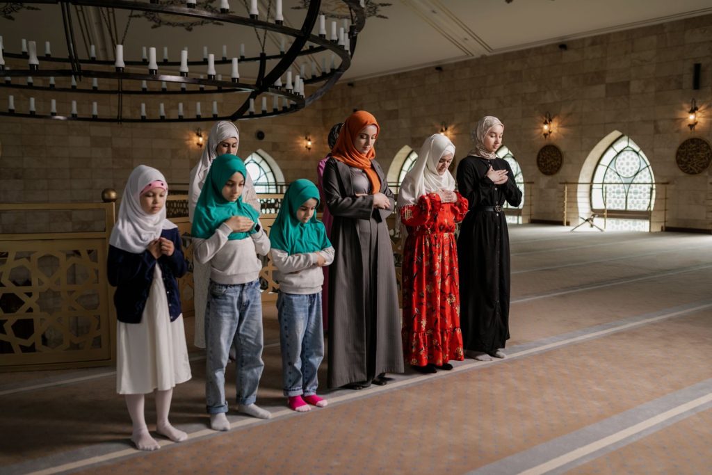 A group of women and children in hijabs praying inside a beautifully lit mosque.
